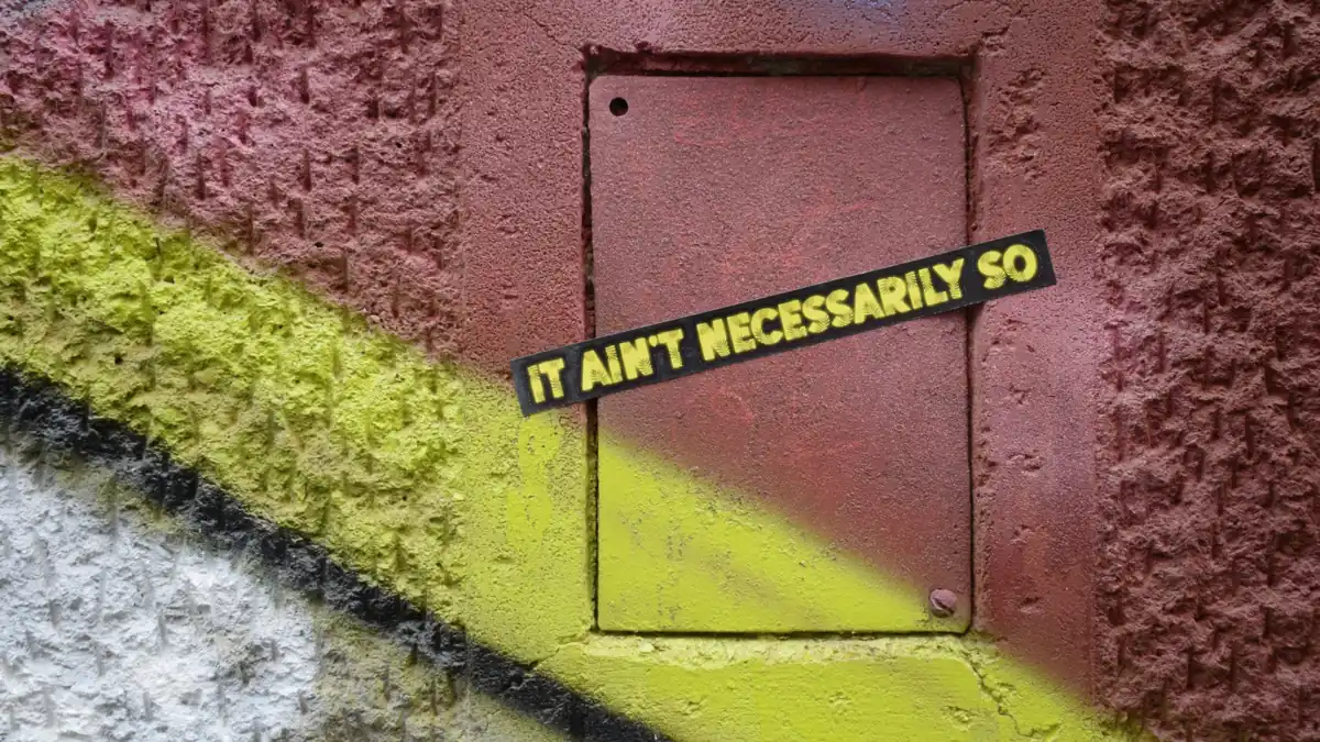 A graffitied cement wall with a vinyl sticker that says “It ain’t necessarily so”